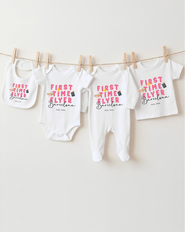 A row of four white baby clothes with the text "FIRST TIME FLYER Barcelona" printed on them, hanging on a clothesline.