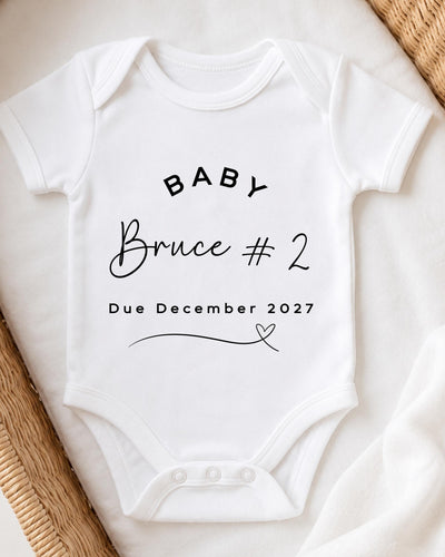 A white baby bodysuit with text printed on it, placed on a bed with a woven blanket.