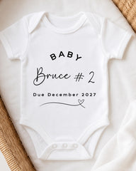 A white baby onesie with text printed on it, placed on a bed with a woven blanket.