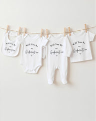 A row of four white baby clothes hanging on a clothesline, each with a different text message printed on it.