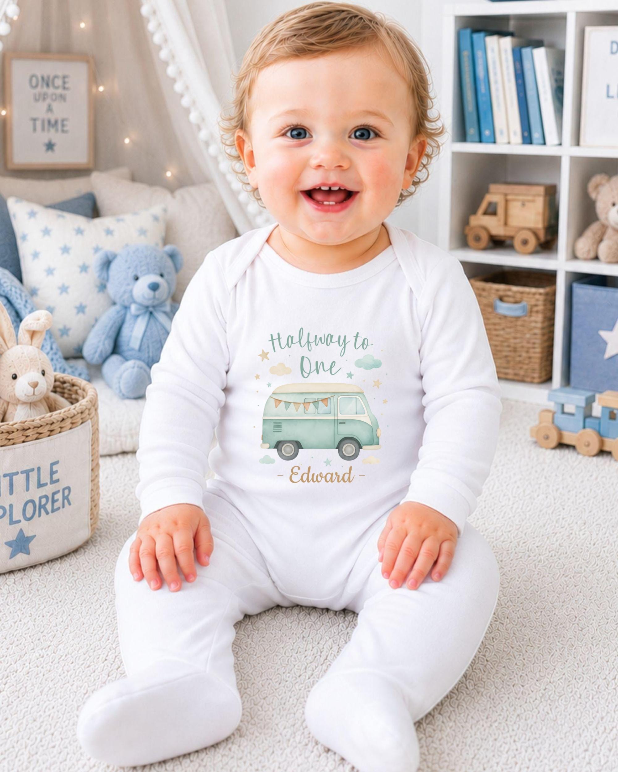 A smiling baby boy wearing a white onesie with a cartoon van design, sitting on a carpeted floor in a room with bookshelves and stuffed animals.