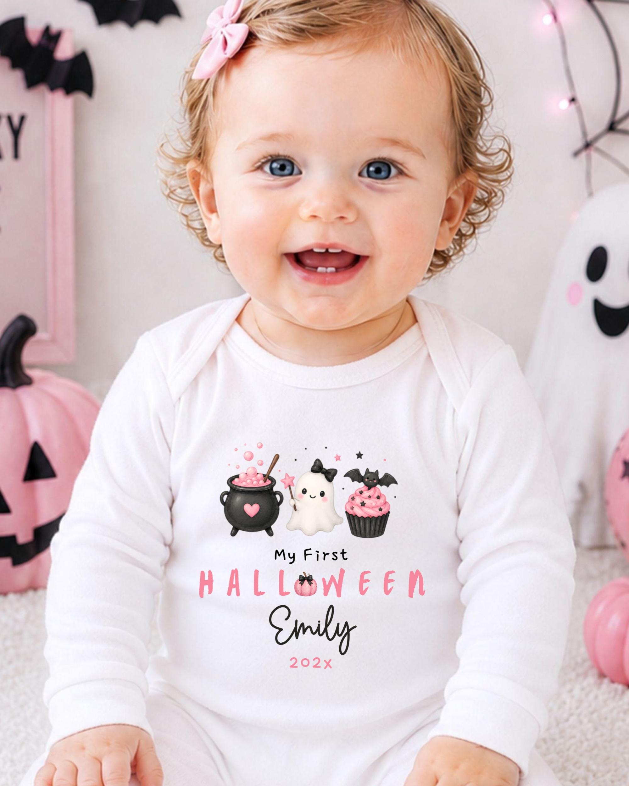 A smiling baby girl wearing a white onesie with a Halloween-themed design, sitting in front of Halloween decorations.