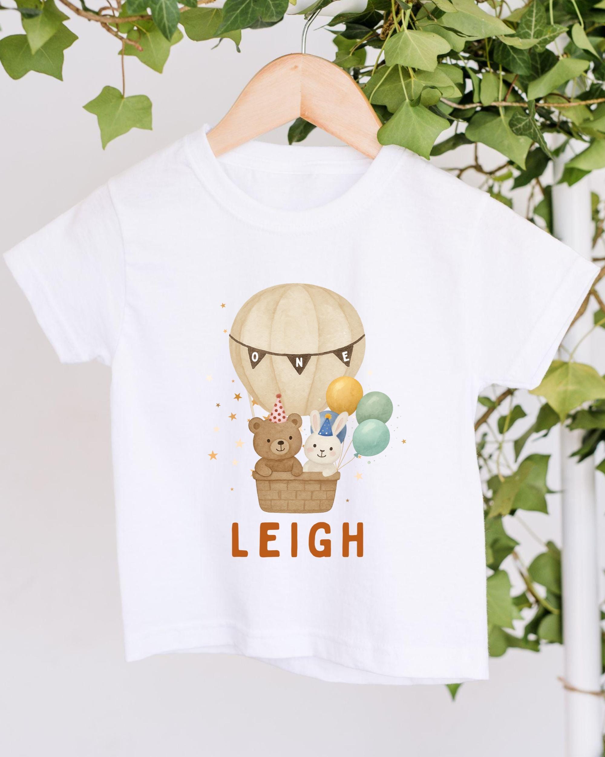 A white t-shirt with a printed design of a hot air balloon carrying a teddy bear and other animals, along with the name "LEIGH" printed below.
