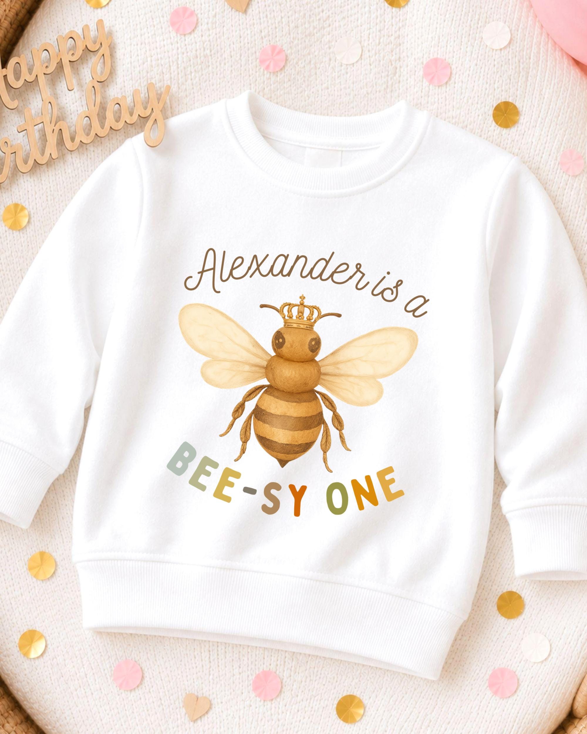 A white sweatshirt with a bee graphic and text printed on it, placed on a surface with confetti.