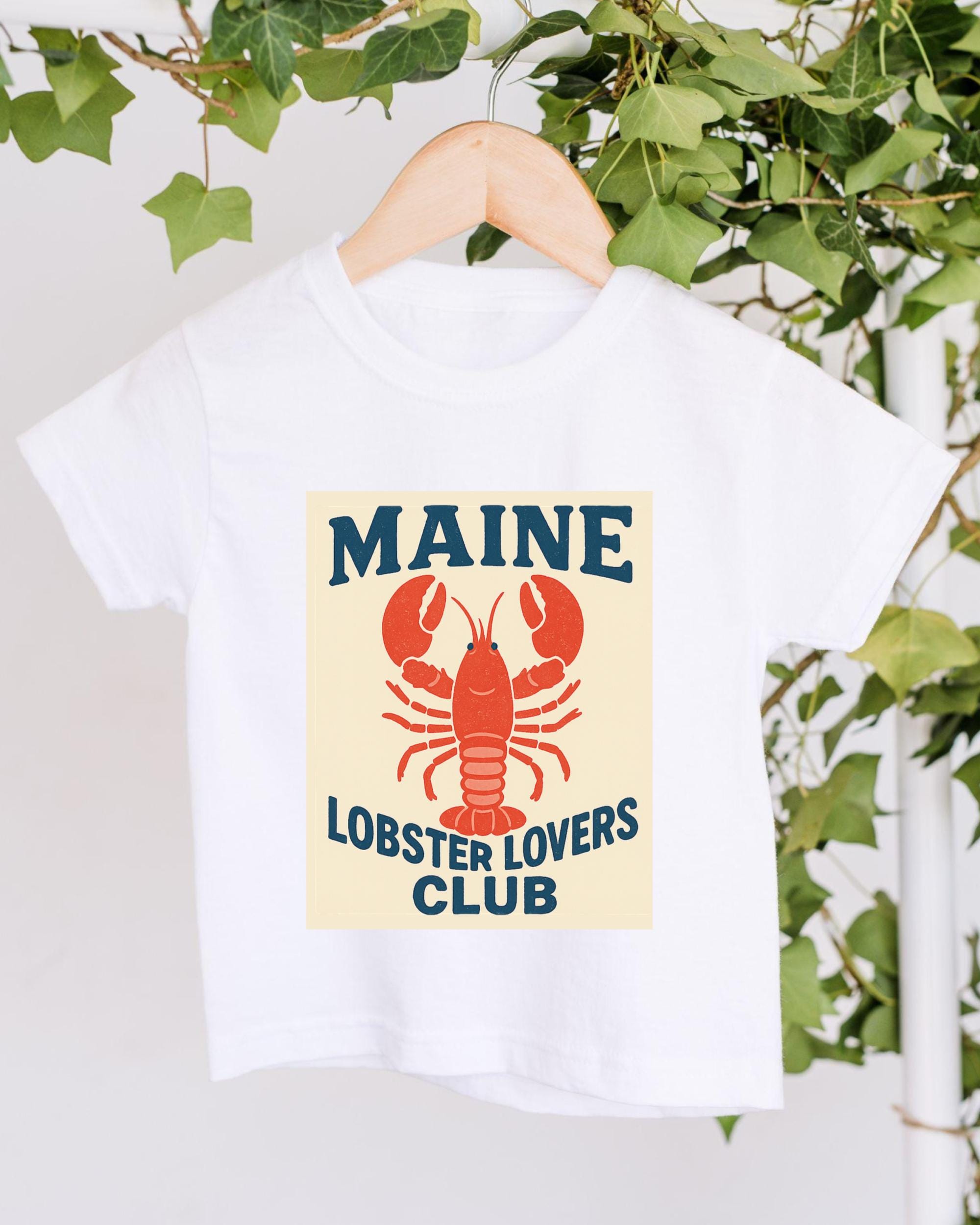 A white t-shirt with a lobster graphic and the text "MAINE LOBSTER LOYALERS CLUB" printed on it, hanging on a wooden hanger.