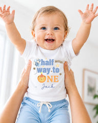 A smiling baby wearing a white onesie with the text "Half Way to One Jack" is being held up by someone's arms.