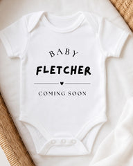 A white baby onesie with the text "BABY FLETCHER COMING SOON" printed on it.
