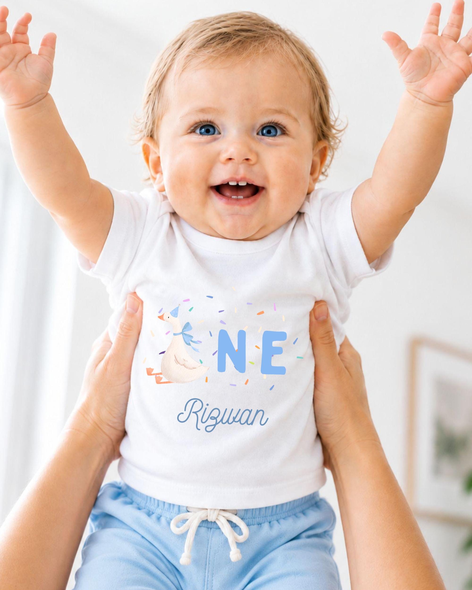 A smiling baby wearing a white shirt with the name "Riquan" and colorful confetti on it, being held up by someone's arms.