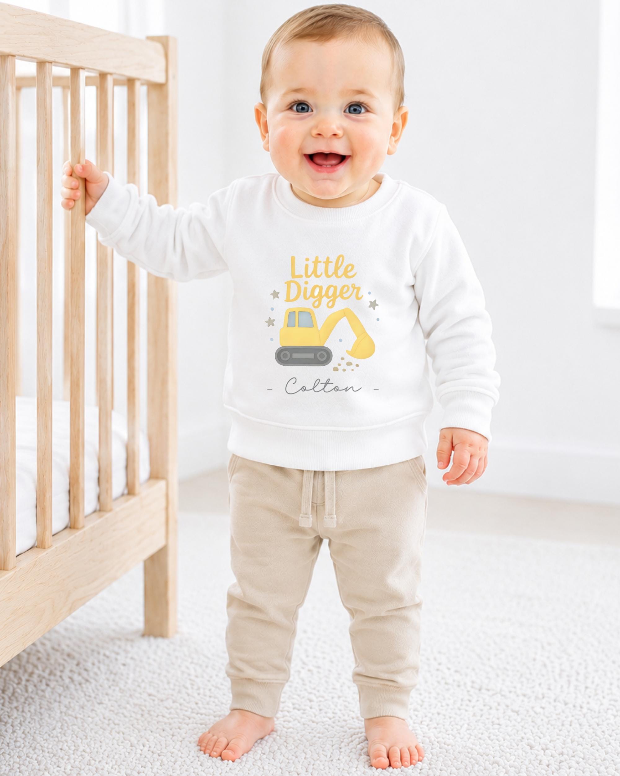 A smiling baby boy wearing a white sweatshirt with a yellow construction vehicle graphic and the text "Little Digger" is standing next to a wooden crib.