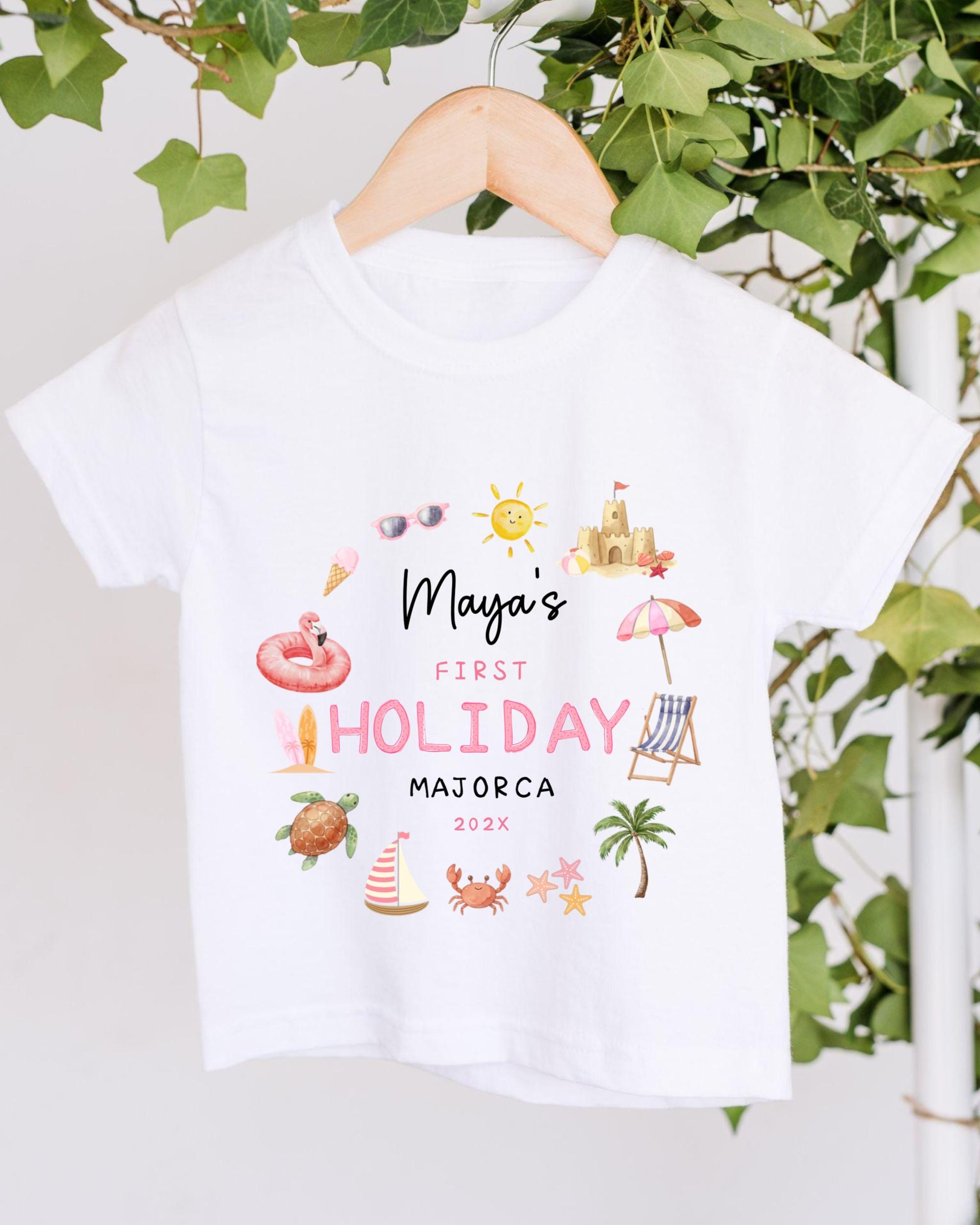 A white t-shirt with a printed design featuring various summer-themed illustrations, including a beach, palm tree, umbrella, and other beach-related objects. The text on the t-shirt reads "Maya's FIRST HOLIDAY MAJORCA 2023".