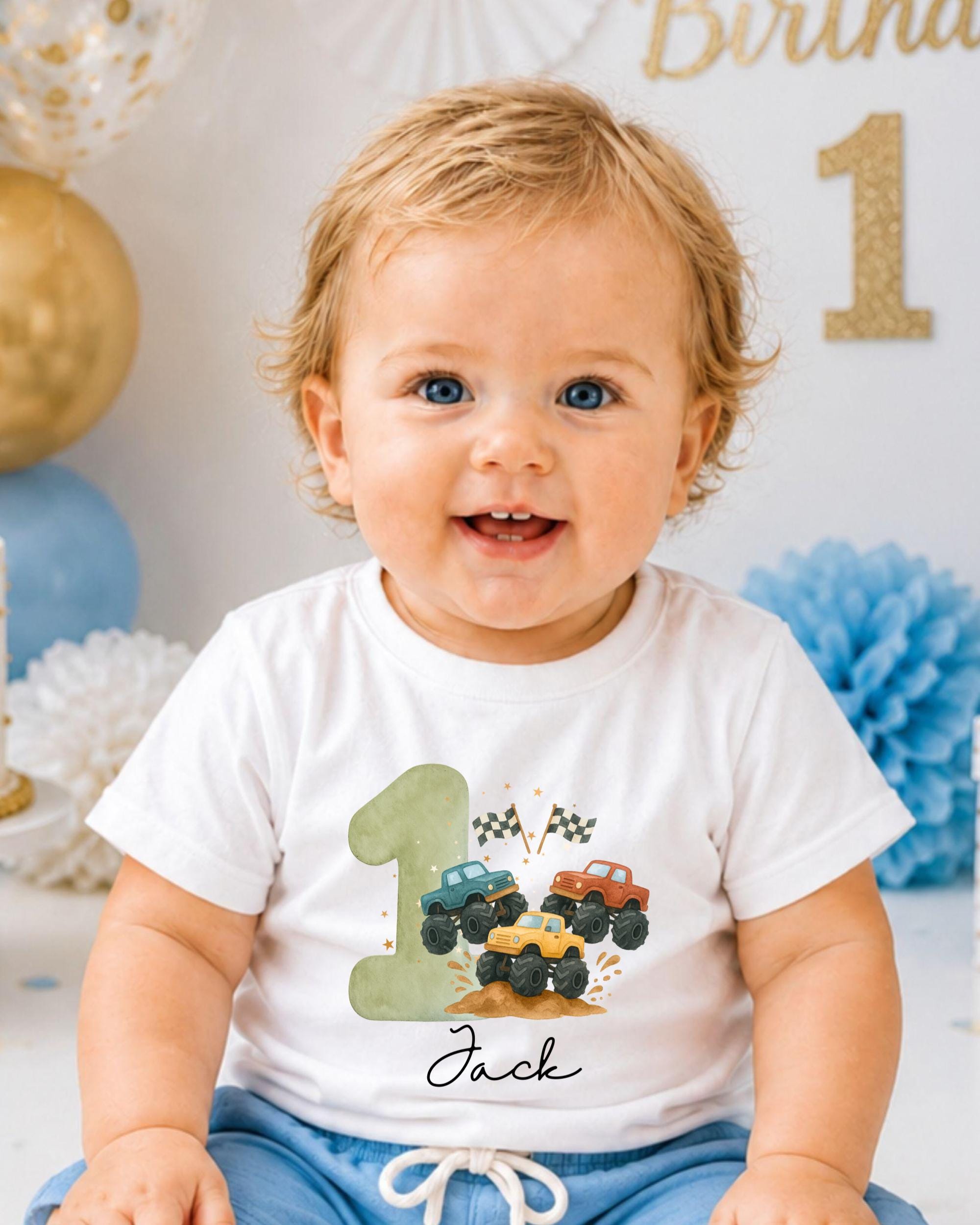 A smiling baby boy wearing a white t-shirt with a graphic of a truck and the number 1, sitting in front of a birthday party backdrop.