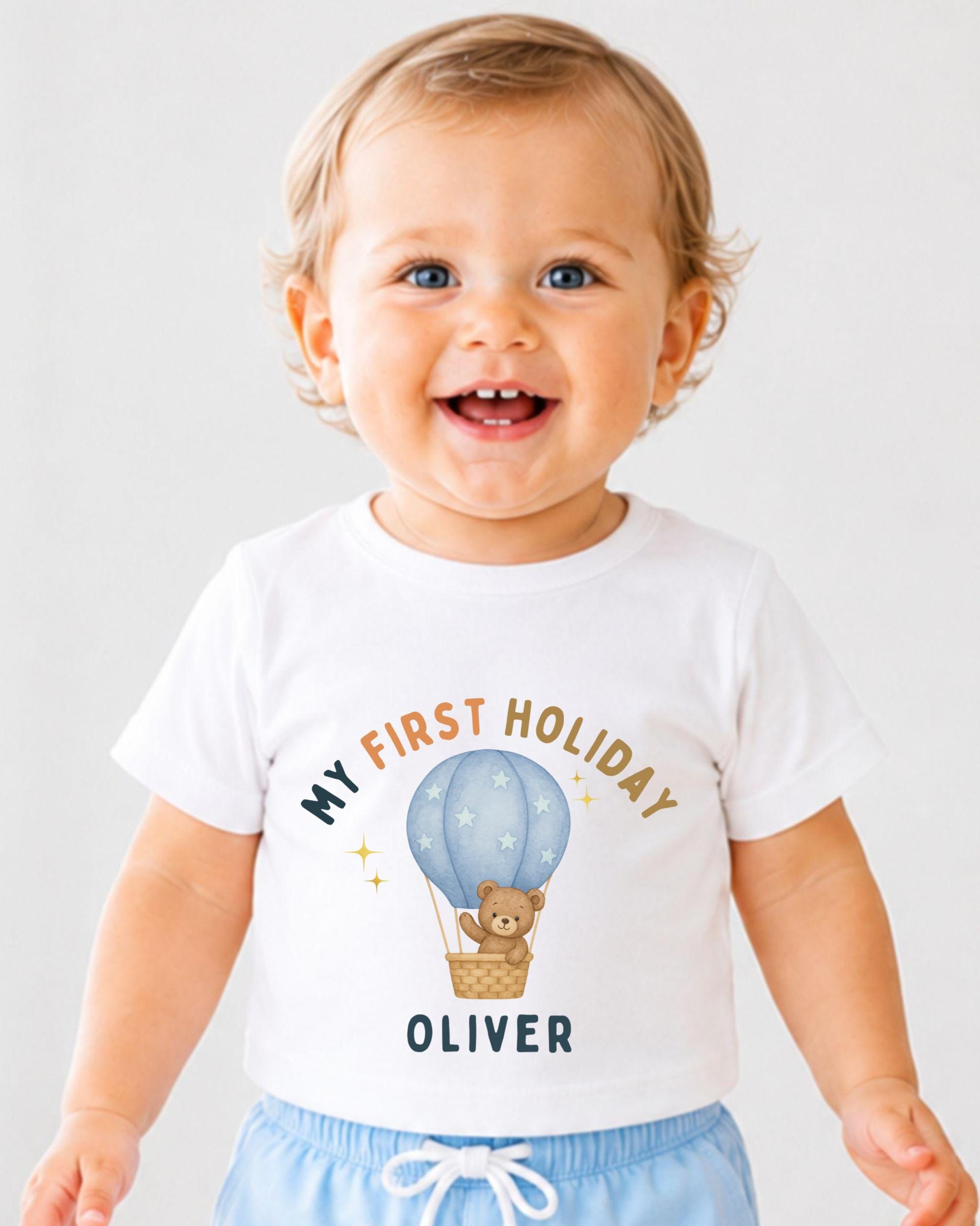 A smiling baby boy wearing a white t-shirt with a cartoon image of a teddy bear in a hot air balloon and the text "My first holiday Oliver" printed on it.