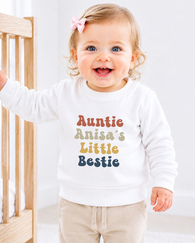 A smiling baby girl wearing a white sweatshirt with the text "Auntie Anisa's Little Bestie" printed on it.