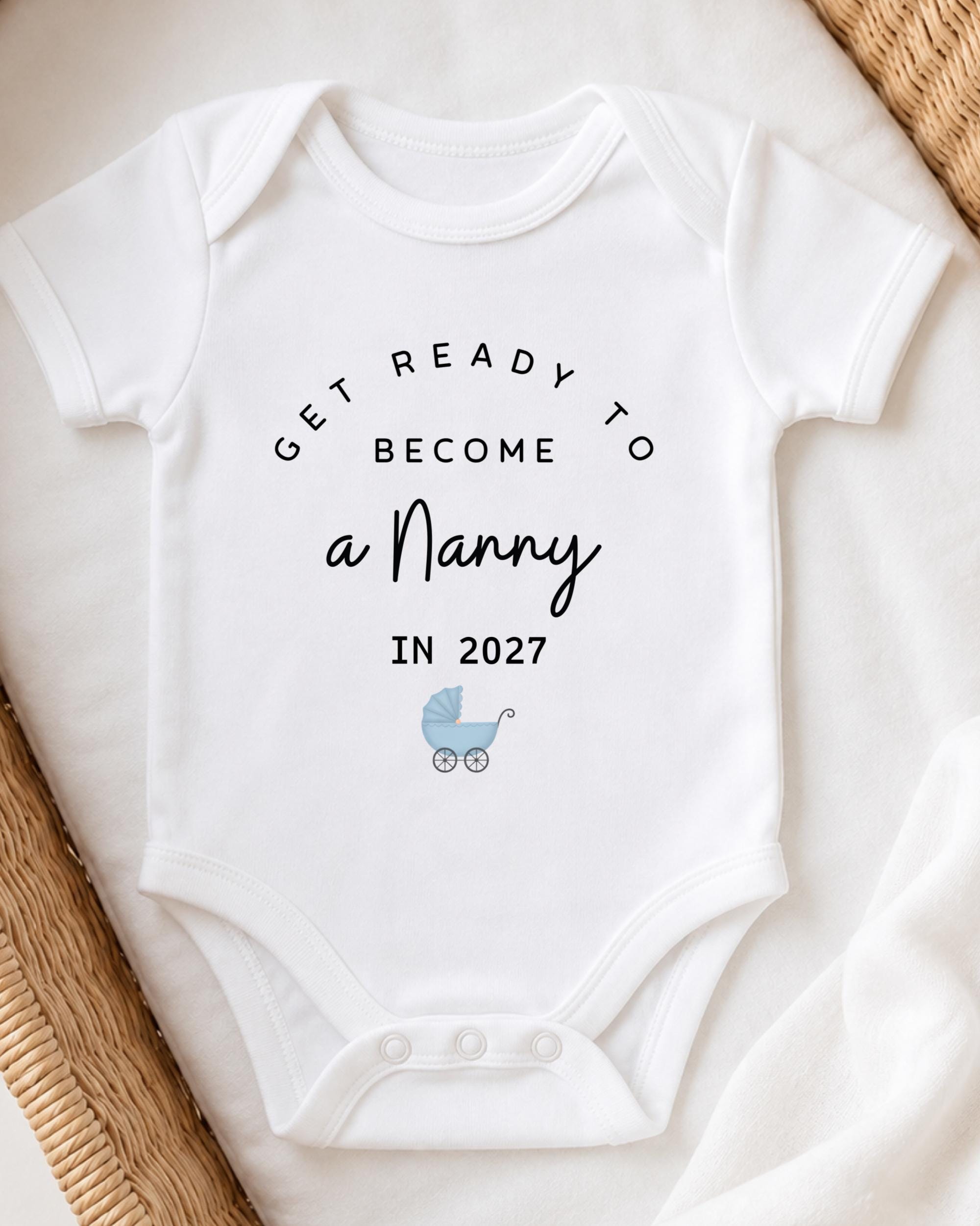 A white onesie with the text "GET READY TO BECOME A NANNY IN 2027" printed on it, along with a small baby carriage icon.