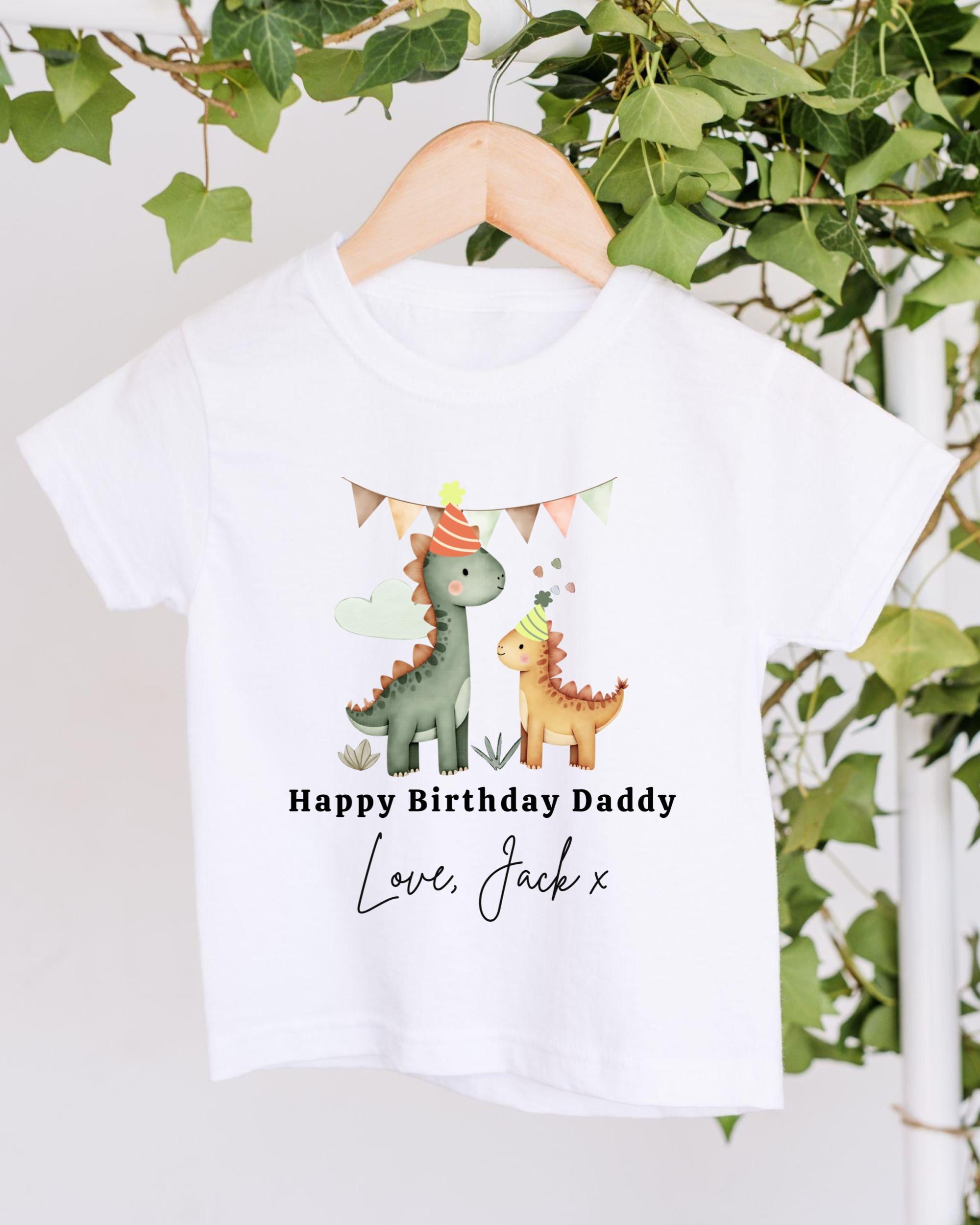 A white t-shirt with a graphic design featuring two cartoon dinosaurs wearing party hats, along with the text "Happy Birthday Daddy Love, Jack x" printed on it.
