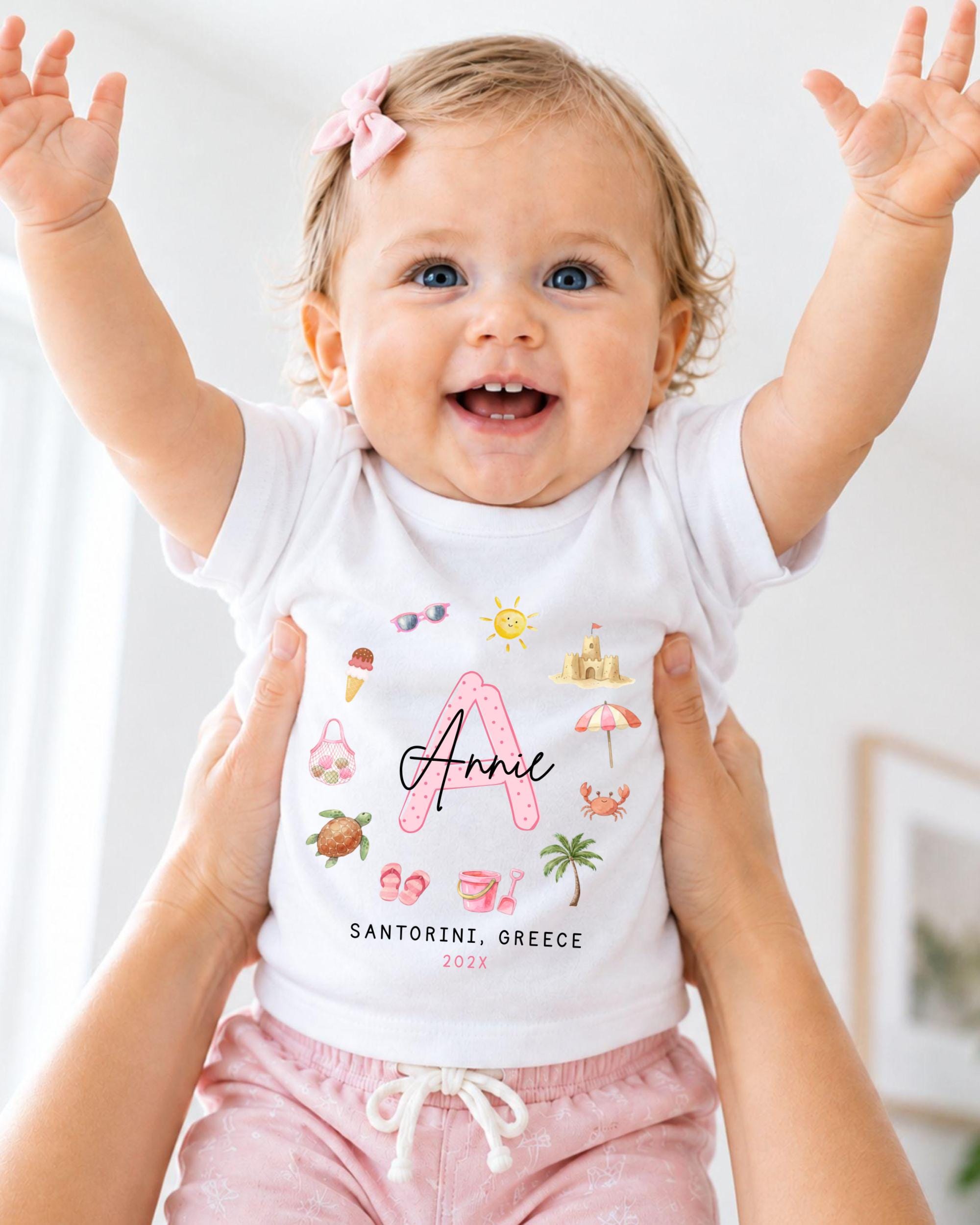 A smiling baby girl wearing a white t-shirt with a personalized name and a beach-themed design, being held up by someone's arms.