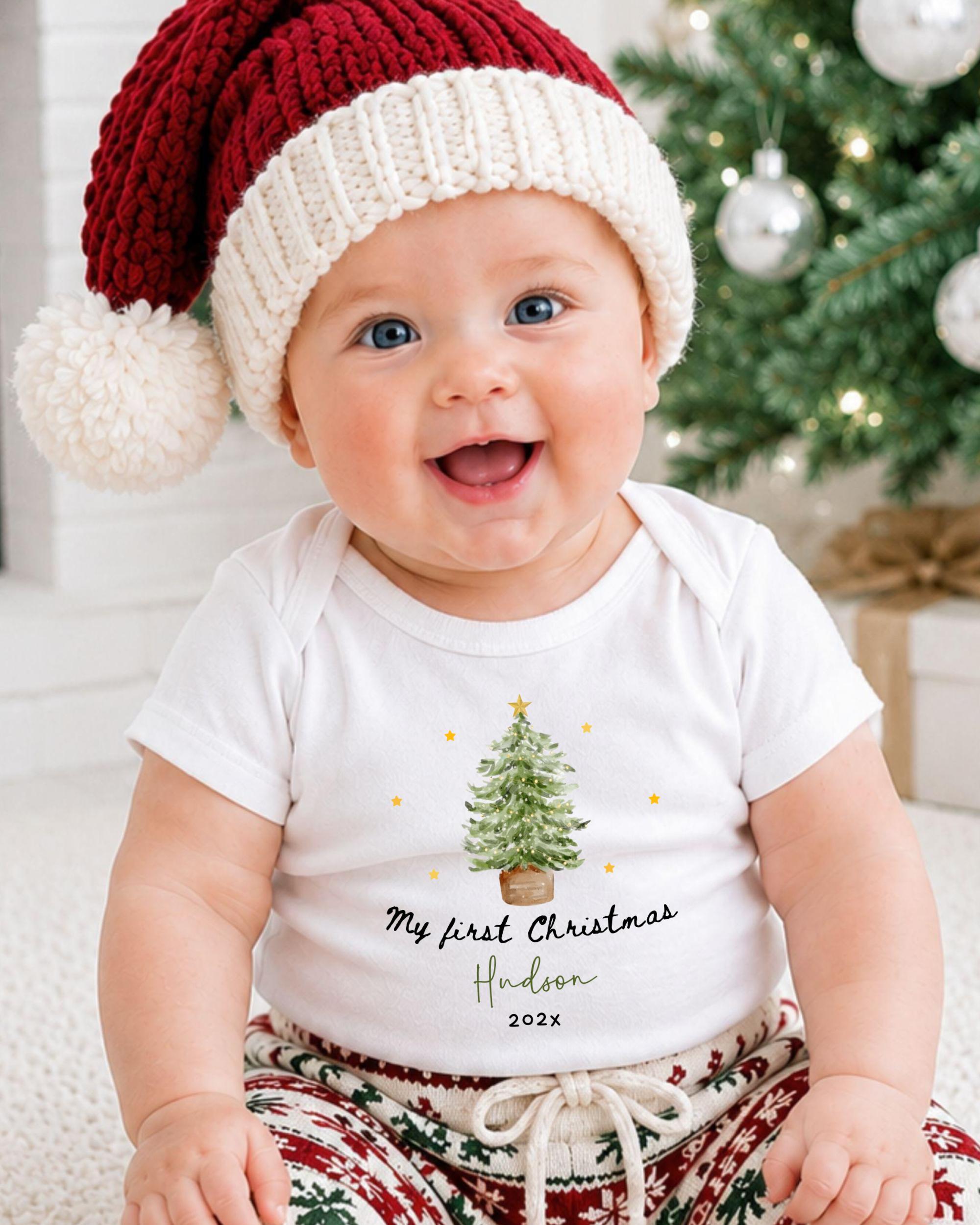 A smiling baby wearing a red and white Santa hat and a white t-shirt with a Christmas tree design, sitting in front of a Christmas tree.