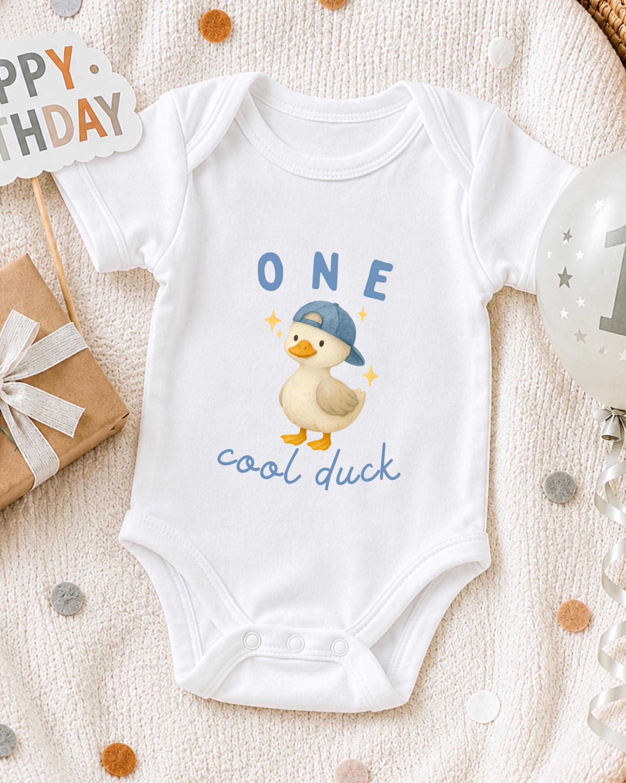 A white onesie with a cartoon duck and the text "One cool duck" printed on it, along with a "Happy Birthday" sign and a balloon.