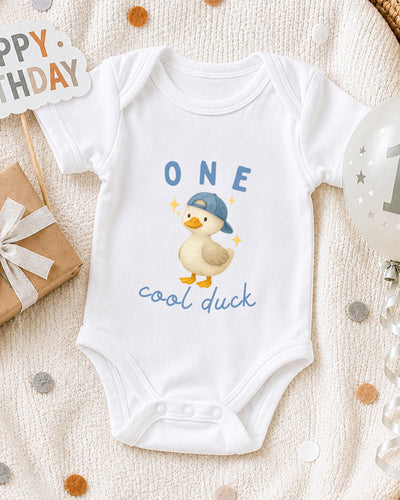 A white bodysuit with a cartoon duck and the text "One cool duck" printed on it, along with a "Happy Birthday" sign and a balloon.