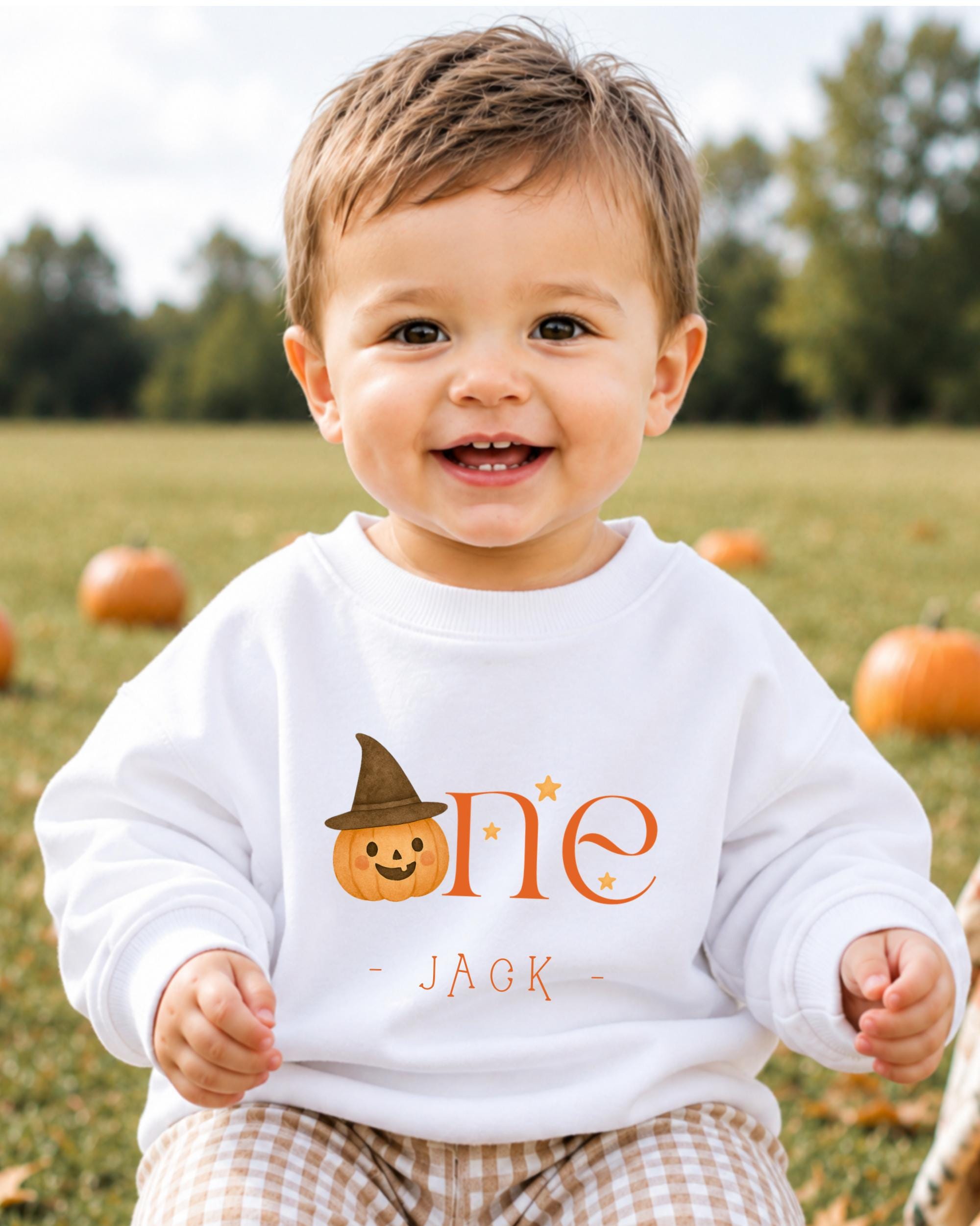 A smiling baby boy wearing a white sweatshirt with a pumpkin and a witch hat, sitting in a field with pumpkins in the background.