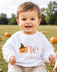 A smiling baby boy wearing a white sweatshirt with a pumpkin and a witch hat, sitting in a field with pumpkins in the background.
