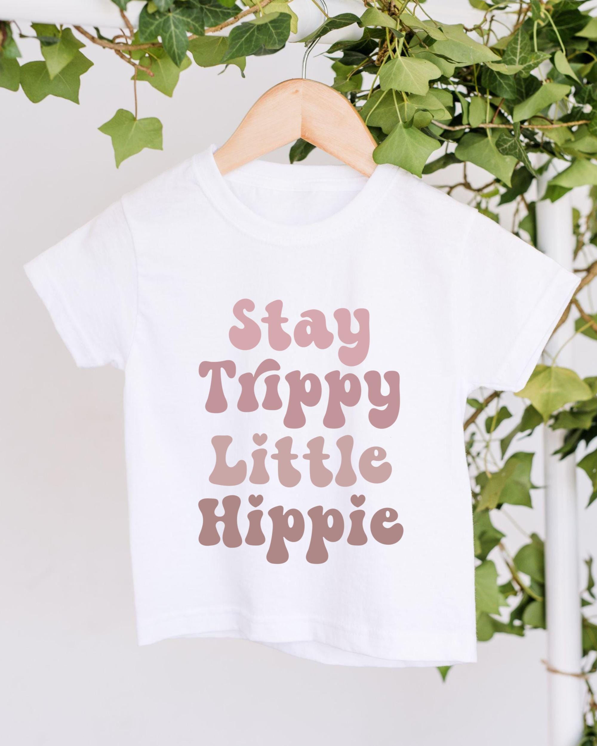 A white t-shirt with the text "Stay Trippy Little Hippie" printed on it, hanging on a wooden hanger.
