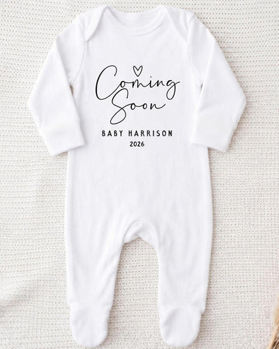 A white bodysuit with the text "Coming Soon" and "Baby Harrison 2026" printed on it.
