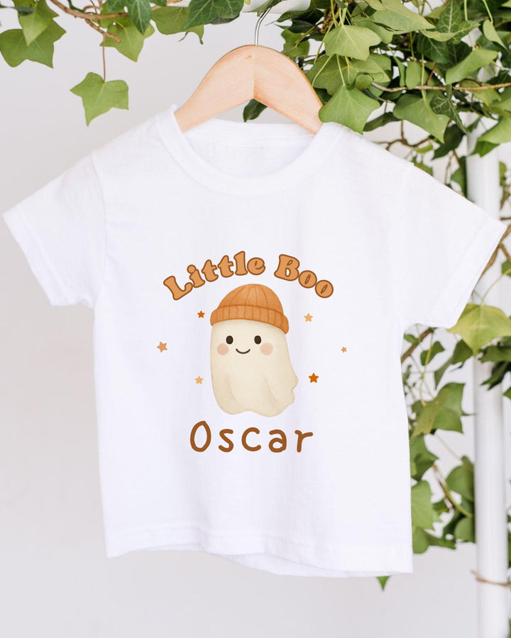 A white t-shirt with a cartoon ghost wearing a hat and the text "Little Boo Oscar" printed on it, hanging on a wooden hanger against a backdrop of greenery.