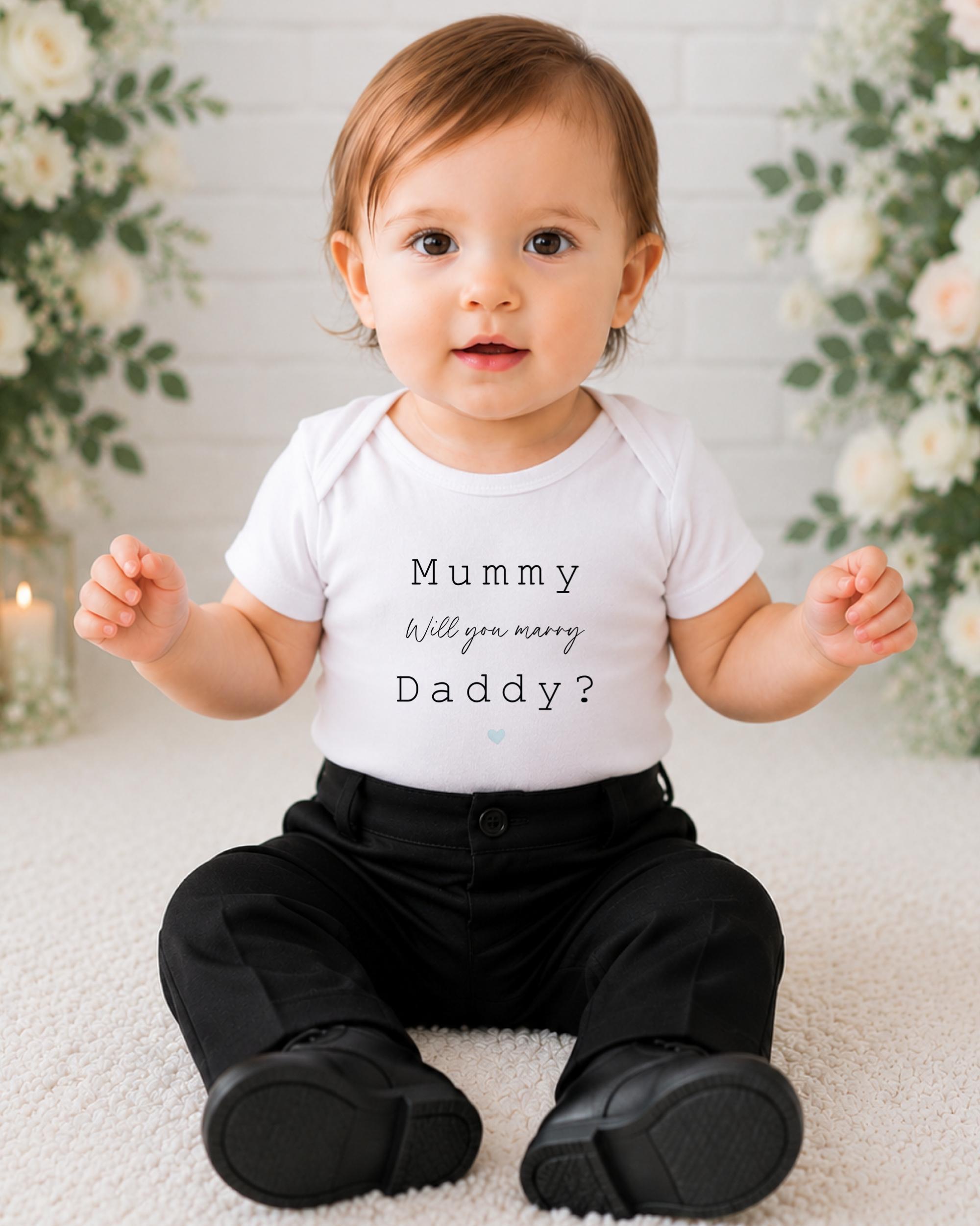 A baby boy wearing a white shirt with the text "Mummy Will you marry Daddy?" and black pants, sitting on a white carpet with flowers in the background.