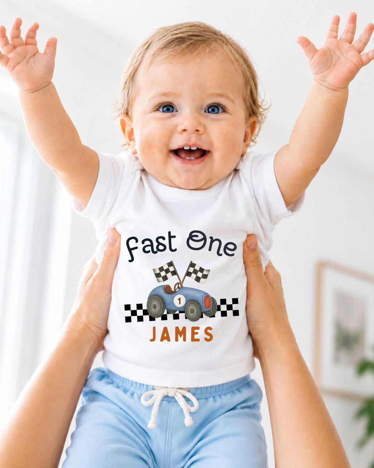 A smiling baby wearing a white t-shirt with a racing car graphic and the name "James" printed on it, being held up by someone's arms.
