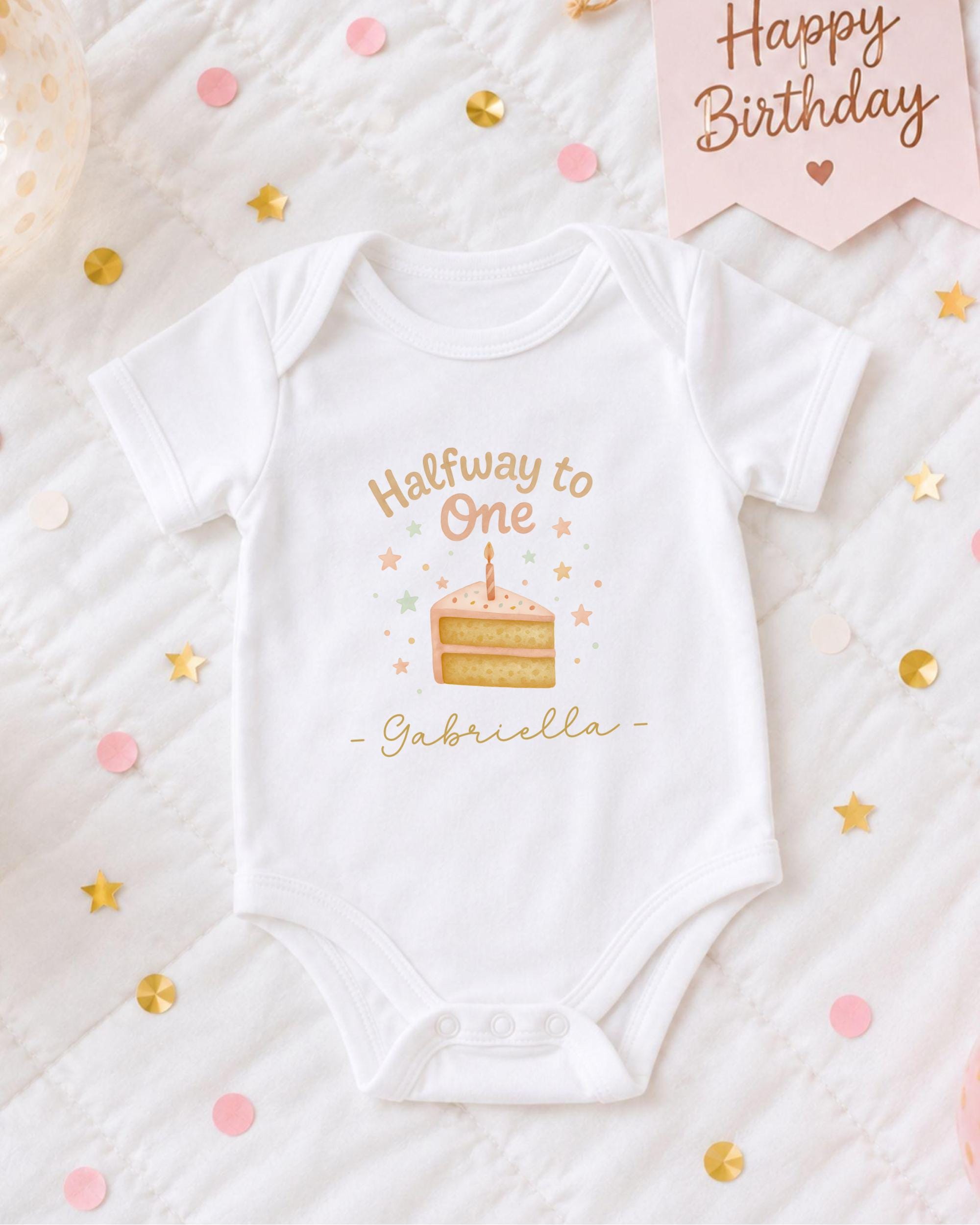 A white onesie with a birthday cake design and the text "Halfway to One Gabriella" printed on it, surrounded by confetti and a "Happy Birthday" sign.