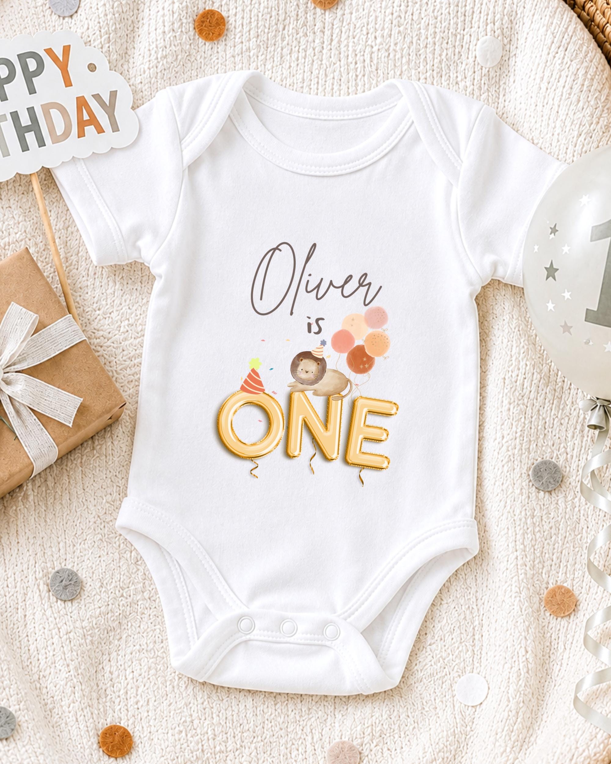 A white onesie with the text "Olivier is 1" printed on it, along with a cartoon image of a lion wearing a party hat. The onesie is placed on a white blanket, surrounded by birthday decorations such as a "Happy Birthday" sign, a balloon, and confetti.