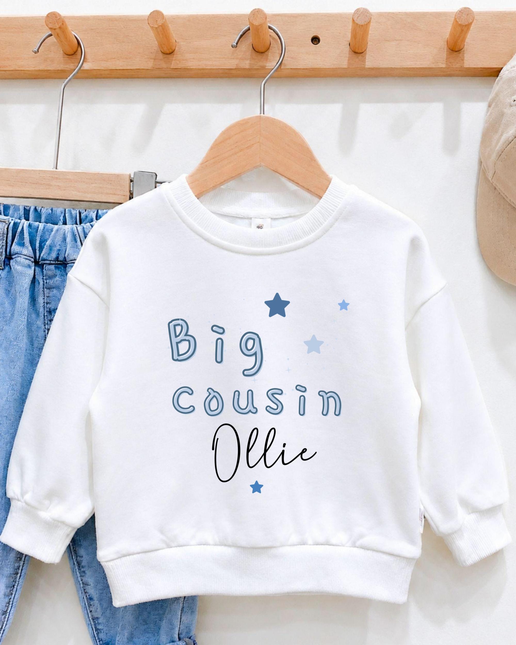 A white sweatshirt with blue stars and text printed on it, hanging on a wooden hanger.