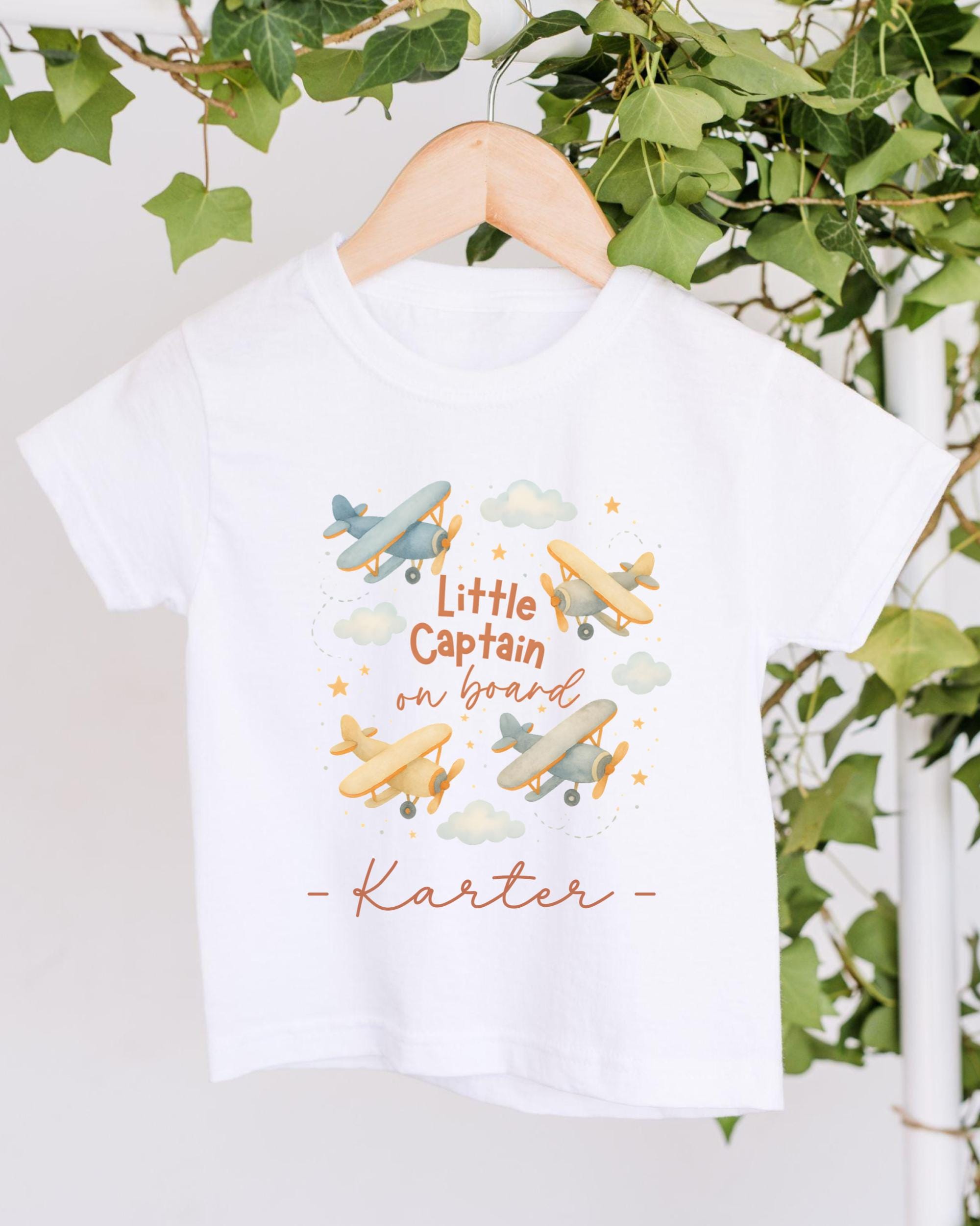 A white t-shirt with a printed design of airplanes and clouds, along with the text "Little Captain on board" and the name "Kater" below it.