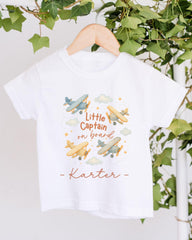 A white t-shirt with a printed design of airplanes and clouds, along with the text "Little Captain on board" and the name "Kater" below it.