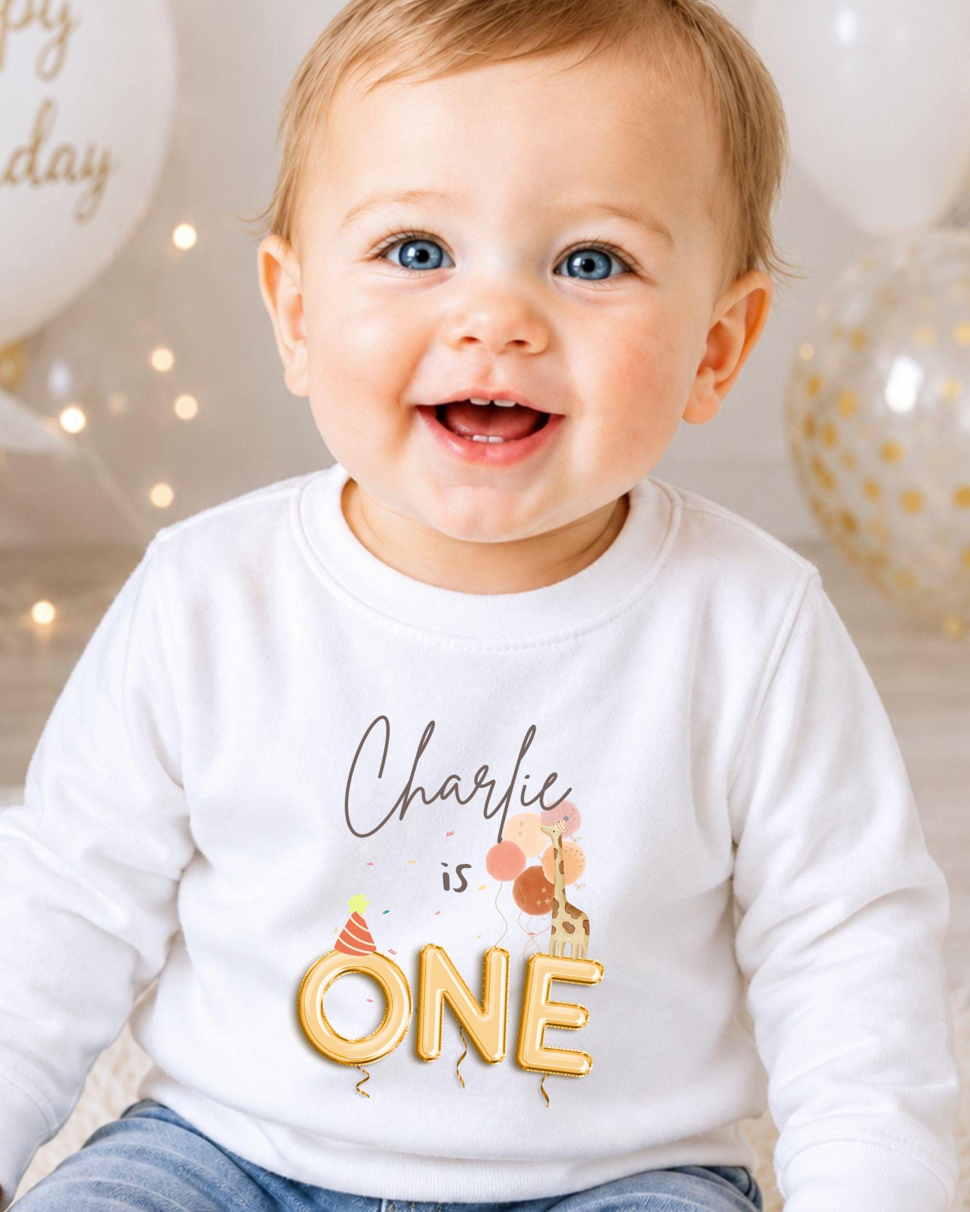 A smiling baby boy wearing a white shirt with the text "Charlie is ONE" printed on it.