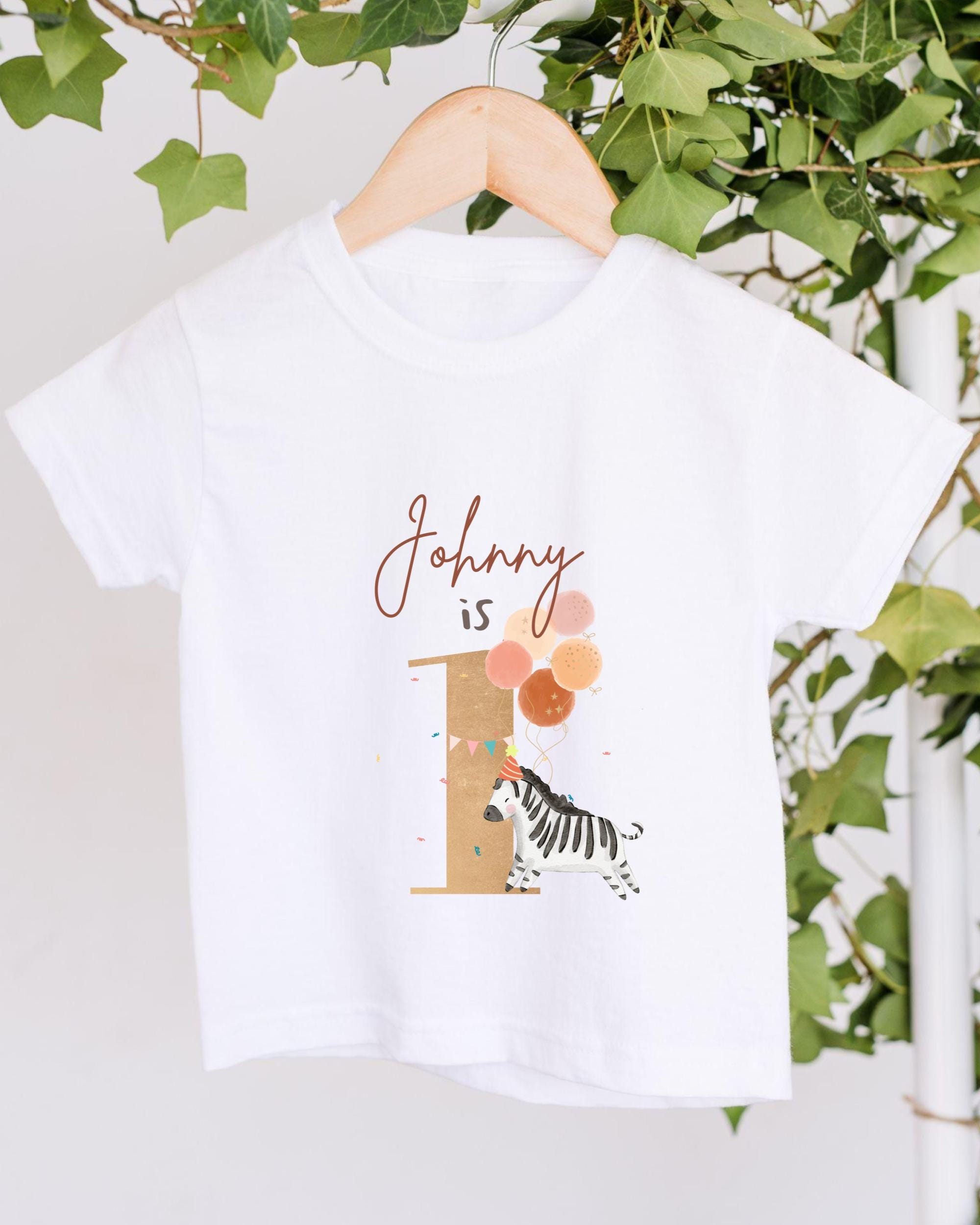 A white t-shirt with a graphic design featuring a zebra and balloons, along with the text "Johny is 1".
