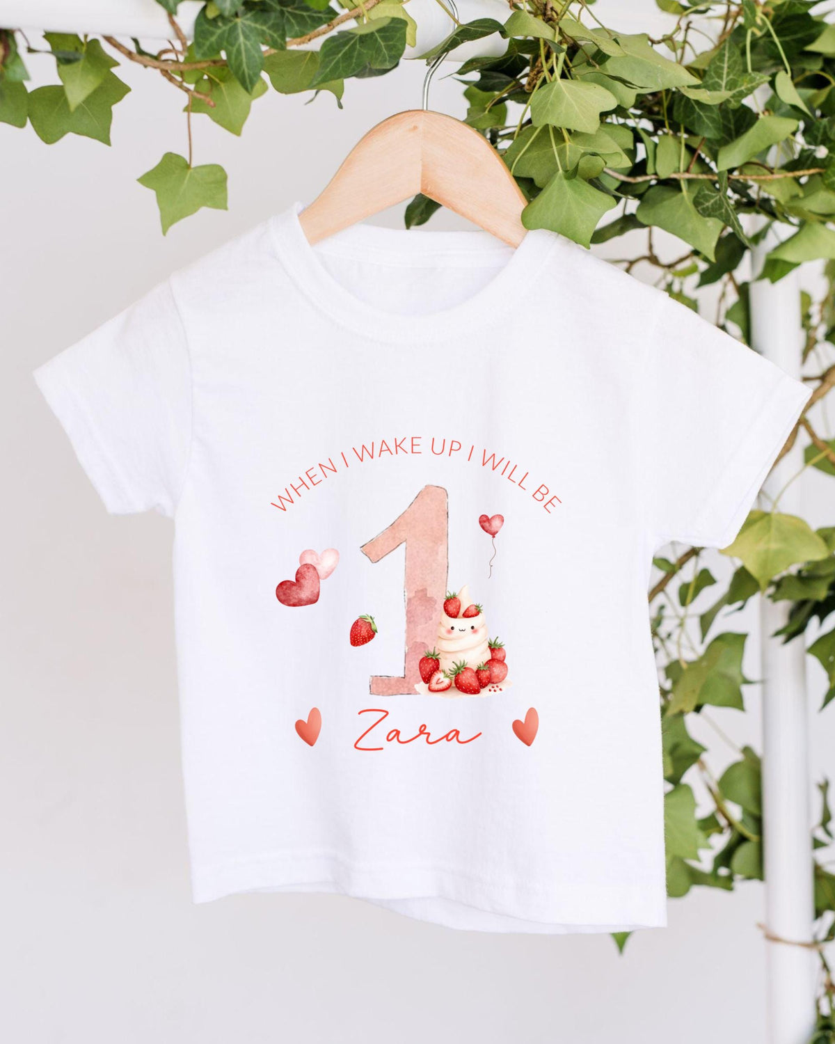 A white t-shirt with a personalized design hanging on a wooden hanger, featuring a pink number one, strawberries, and hearts.