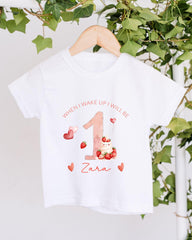 A white t-shirt with a personalized design hanging on a wooden hanger, featuring a pink number one, strawberries, and hearts.