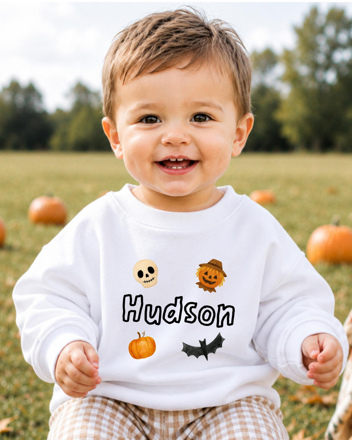 A smiling baby boy wearing a white sweatshirt with a Halloween-themed design, including a skull, pumpkin, bat, and pumpkin.