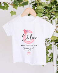 A white t-shirt with a pink floral design and text printed on it, hanging on a wooden hanger.