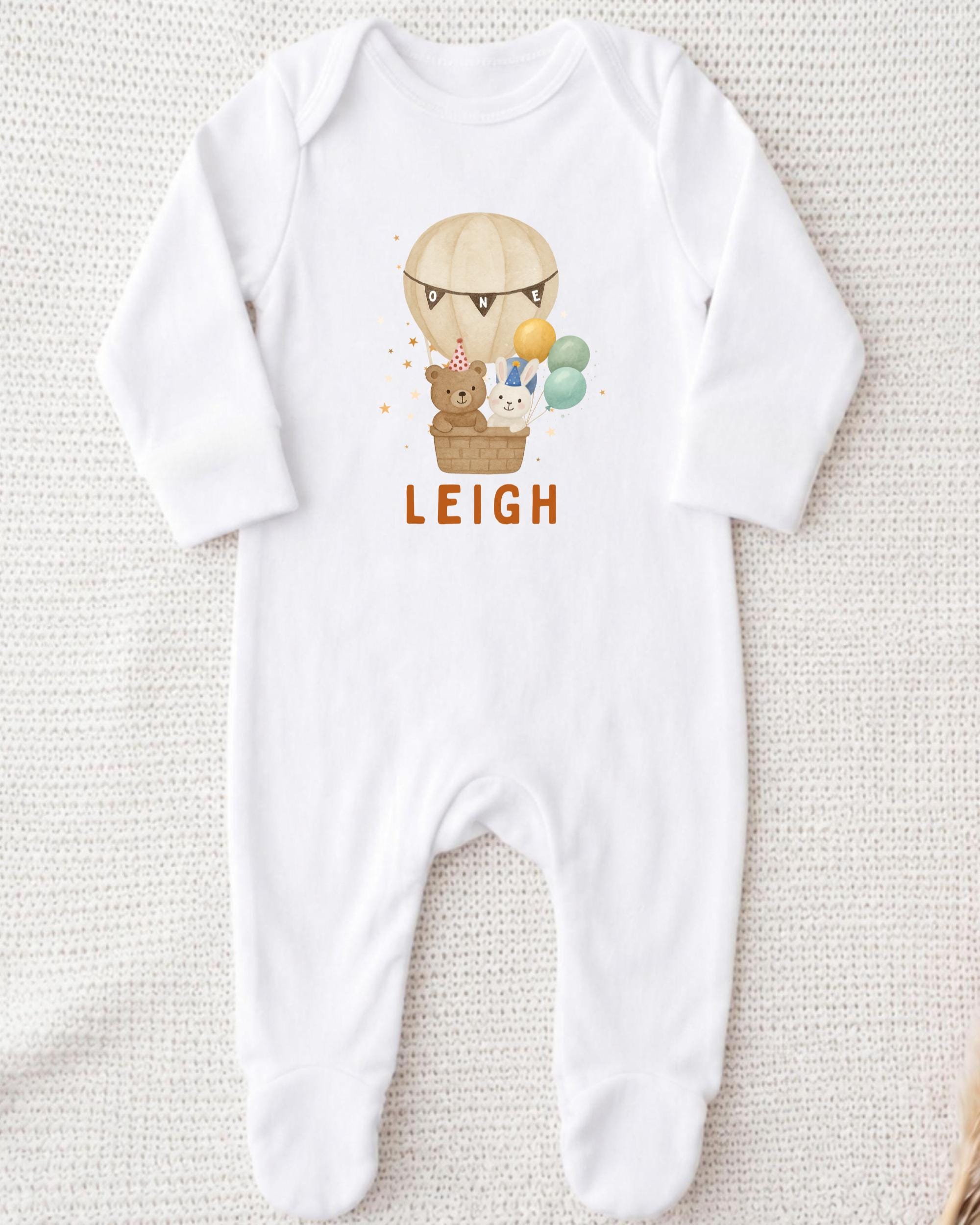 A white onesie with a printed image of a teddy bear and a hot air balloon, along with the name "LEIGH" printed on it.