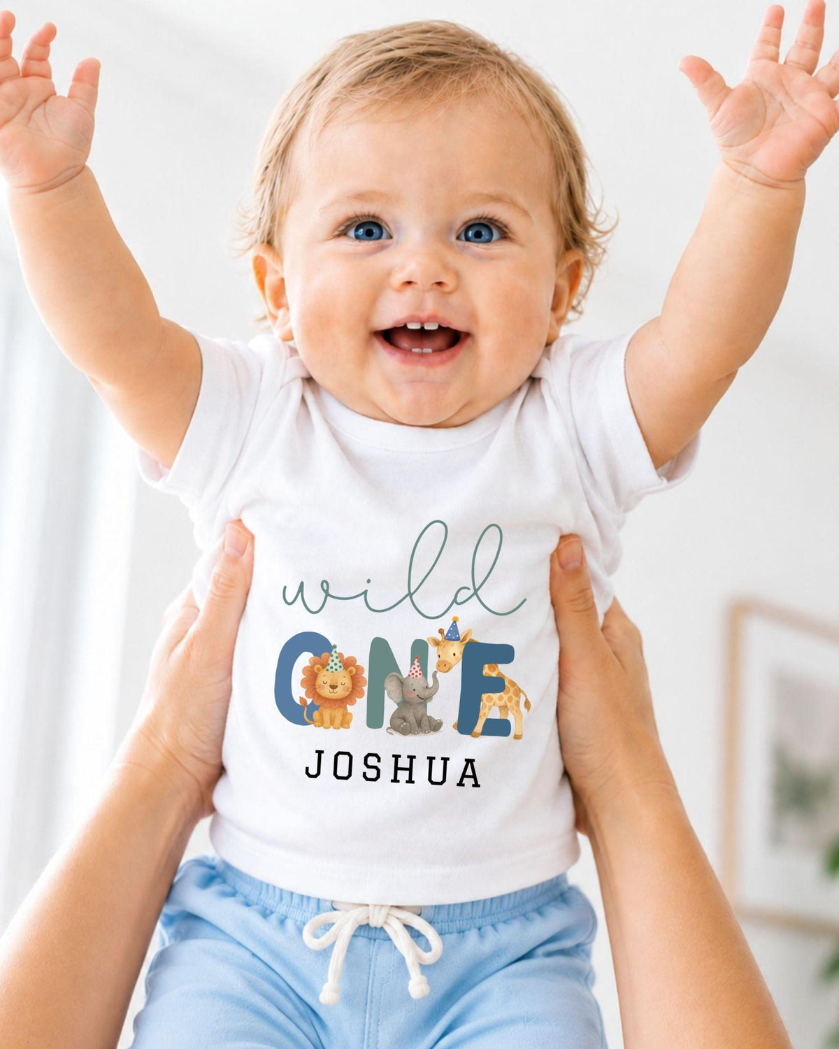 A smiling baby wearing a white t-shirt with a personalized name and animal designs, being held up by someone's arms.
