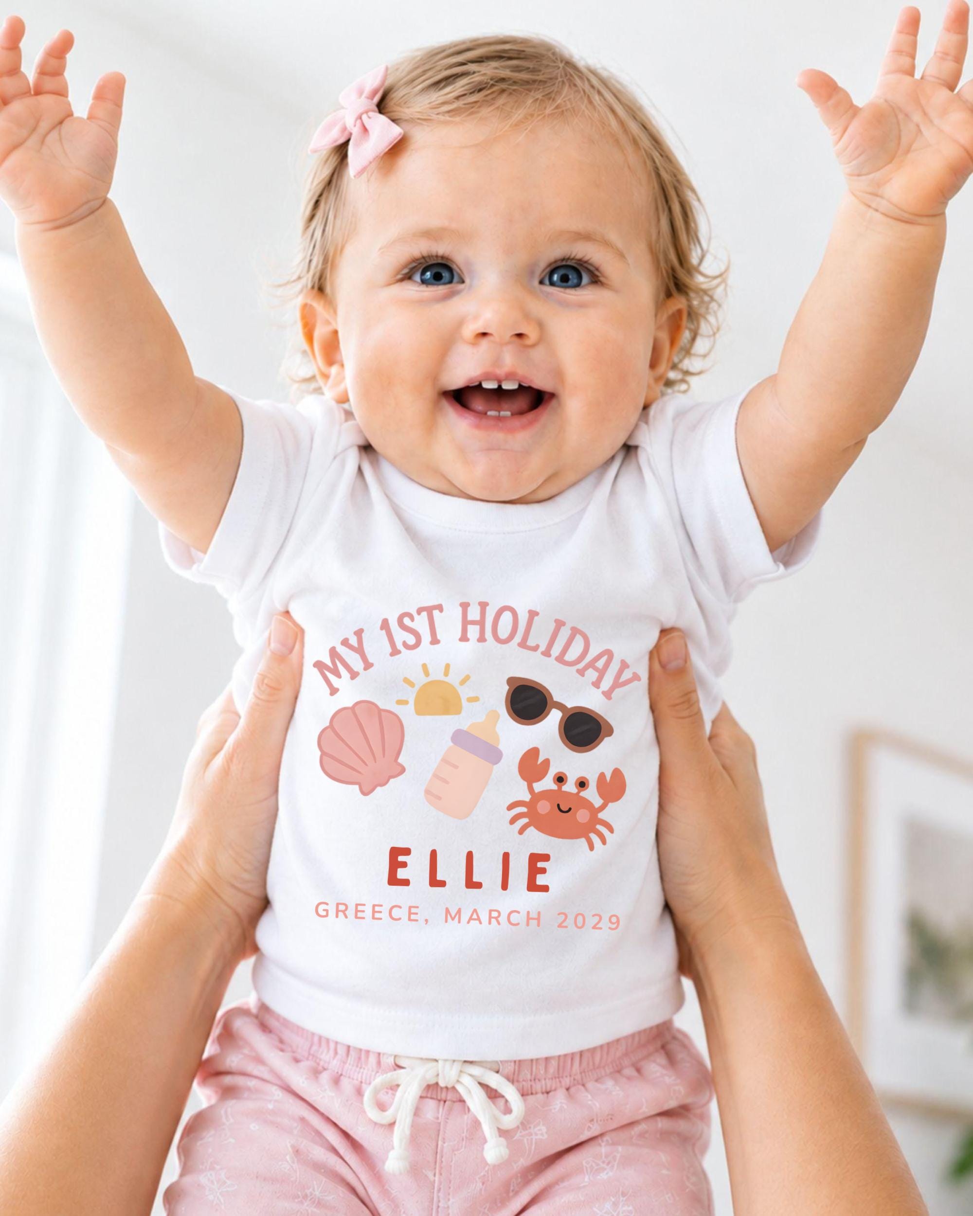 A smiling baby girl wearing a white t-shirt with a graphic design and text, being held up by someone's arms.