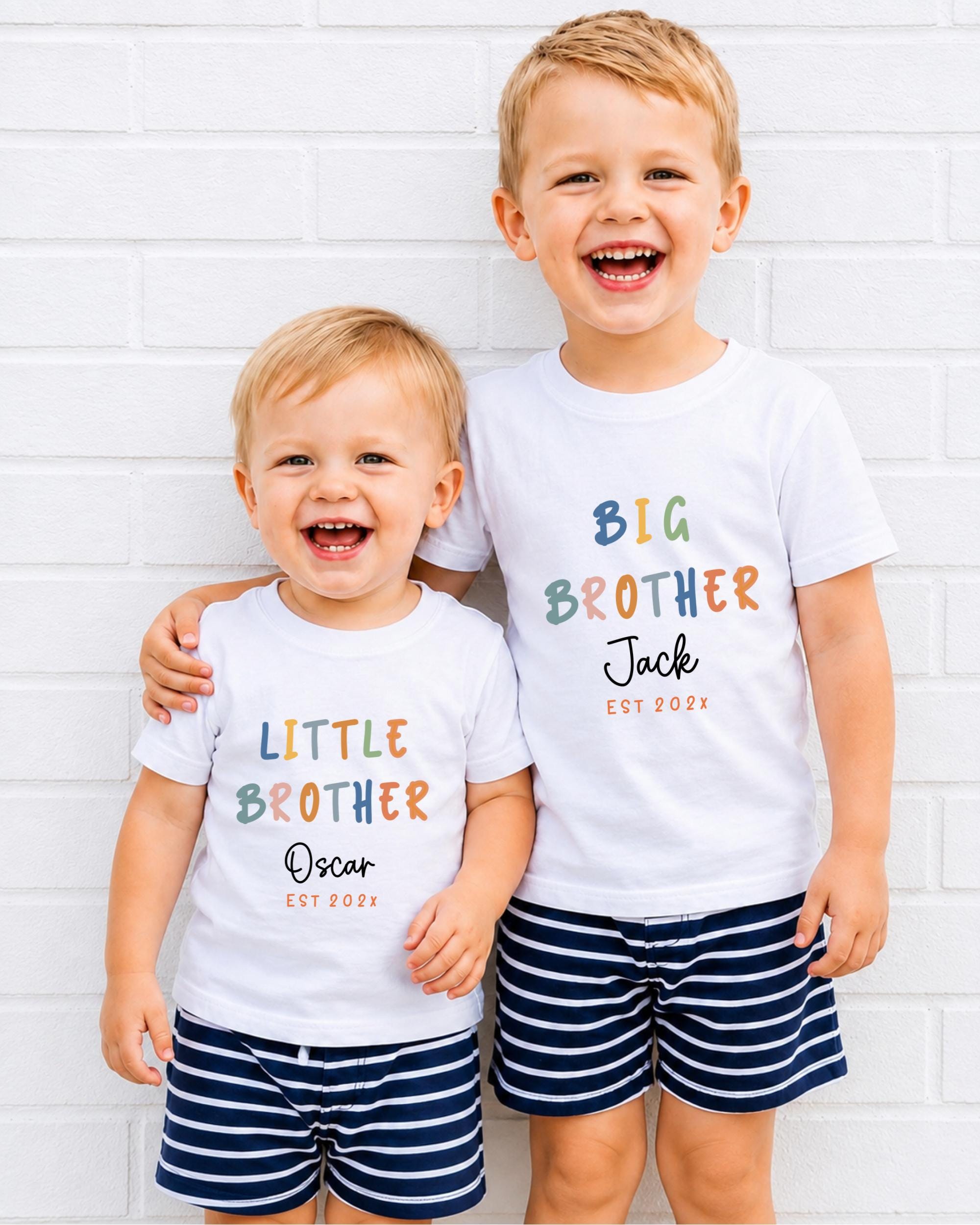 Two young boys wearing matching white t-shirts with text and graphics, smiling and posing together against a white brick wall.