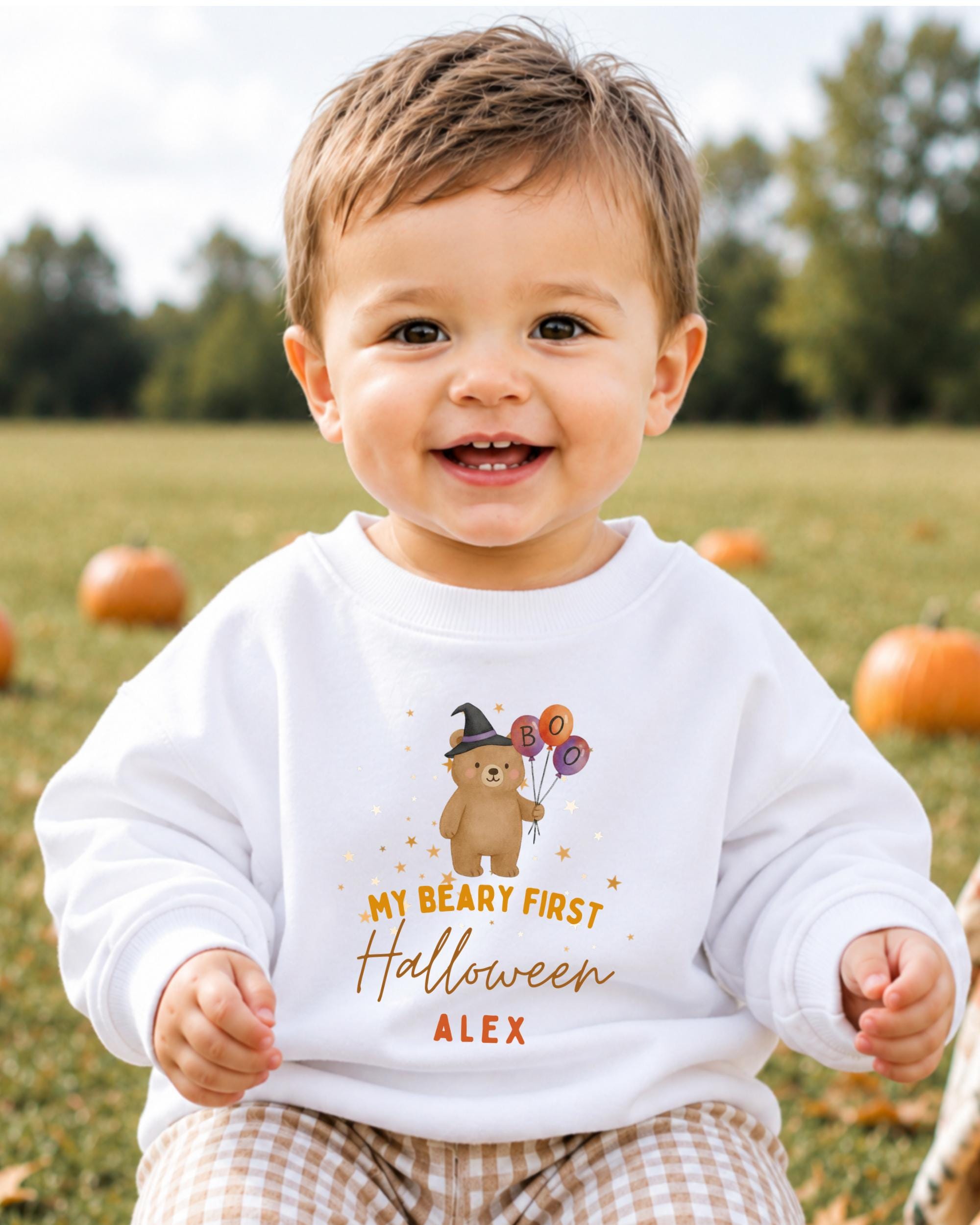 A smiling baby boy wearing a white sweatshirt with a Halloween-themed design and text.