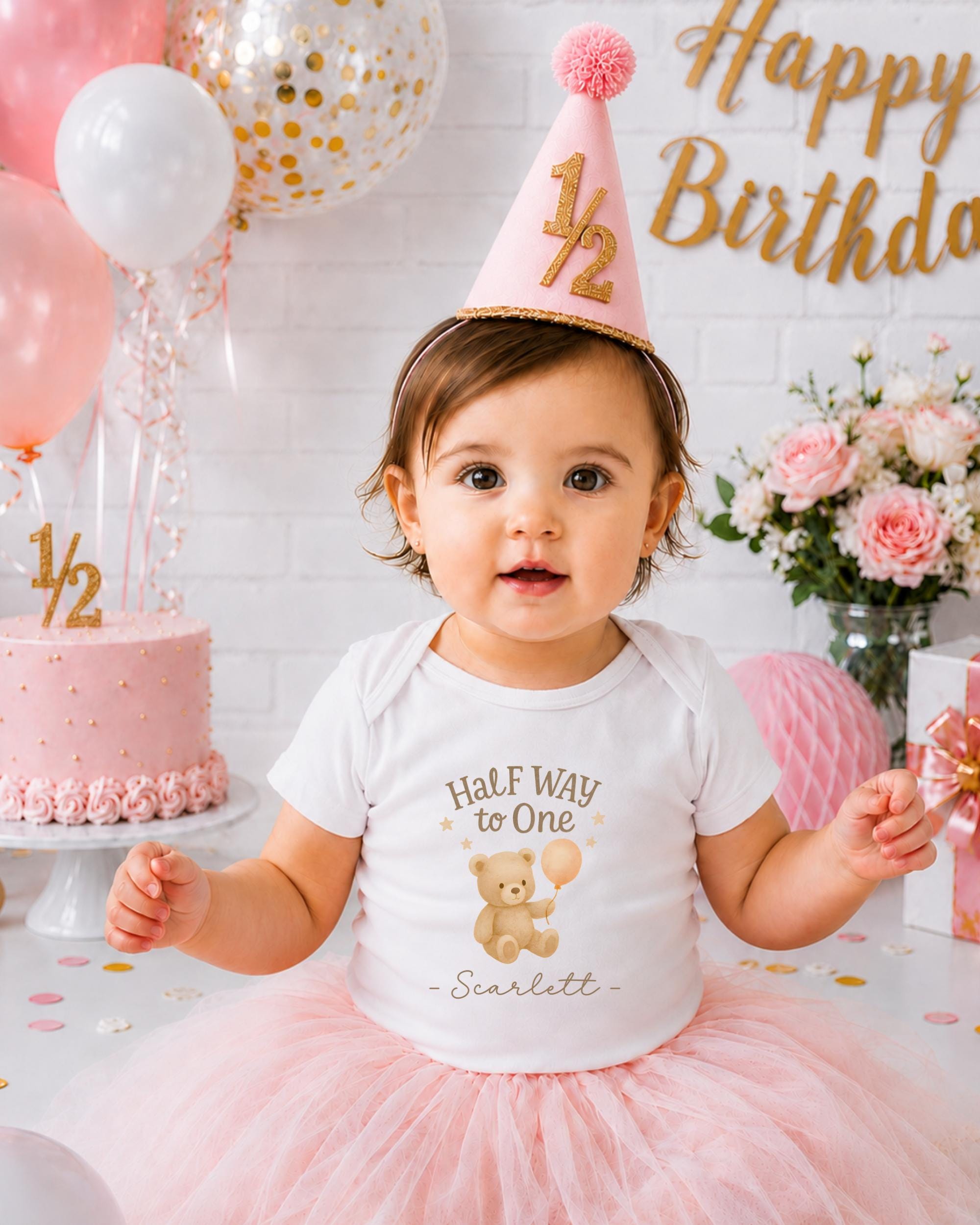 A young girl wearing a white t-shirt with a teddy bear and the text "Half Way to One" is sitting in front of a birthday cake and balloons, with a "Happy Birthday" sign in the background.