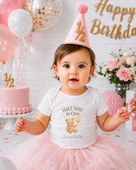 A young girl wearing a white t-shirt with a teddy bear and the text "Half Way to One" is sitting in front of a birthday cake and balloons, with a "Happy Birthday" sign in the background.
