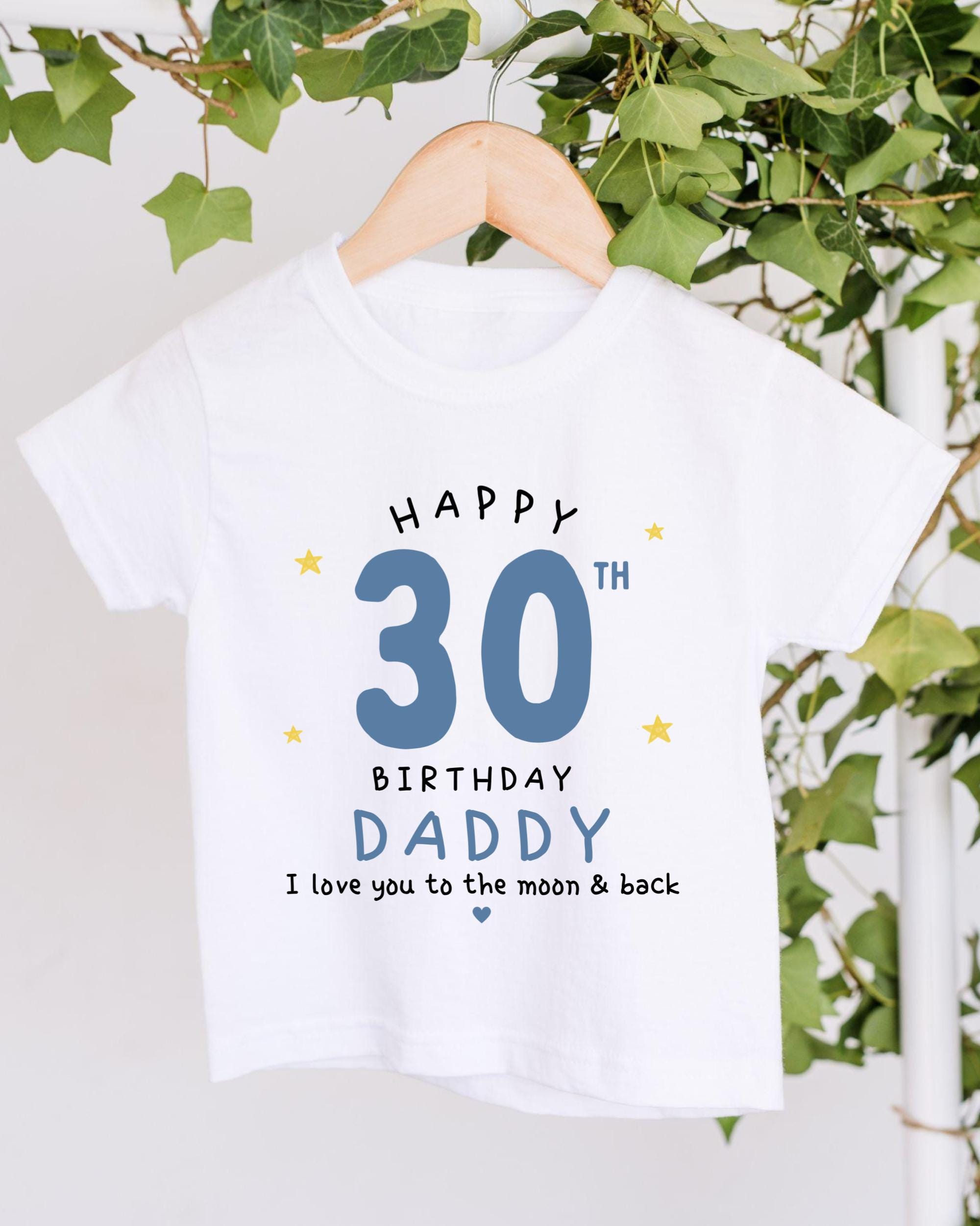 A white t-shirt with a birthday message printed on it, hanging on a wooden hanger.