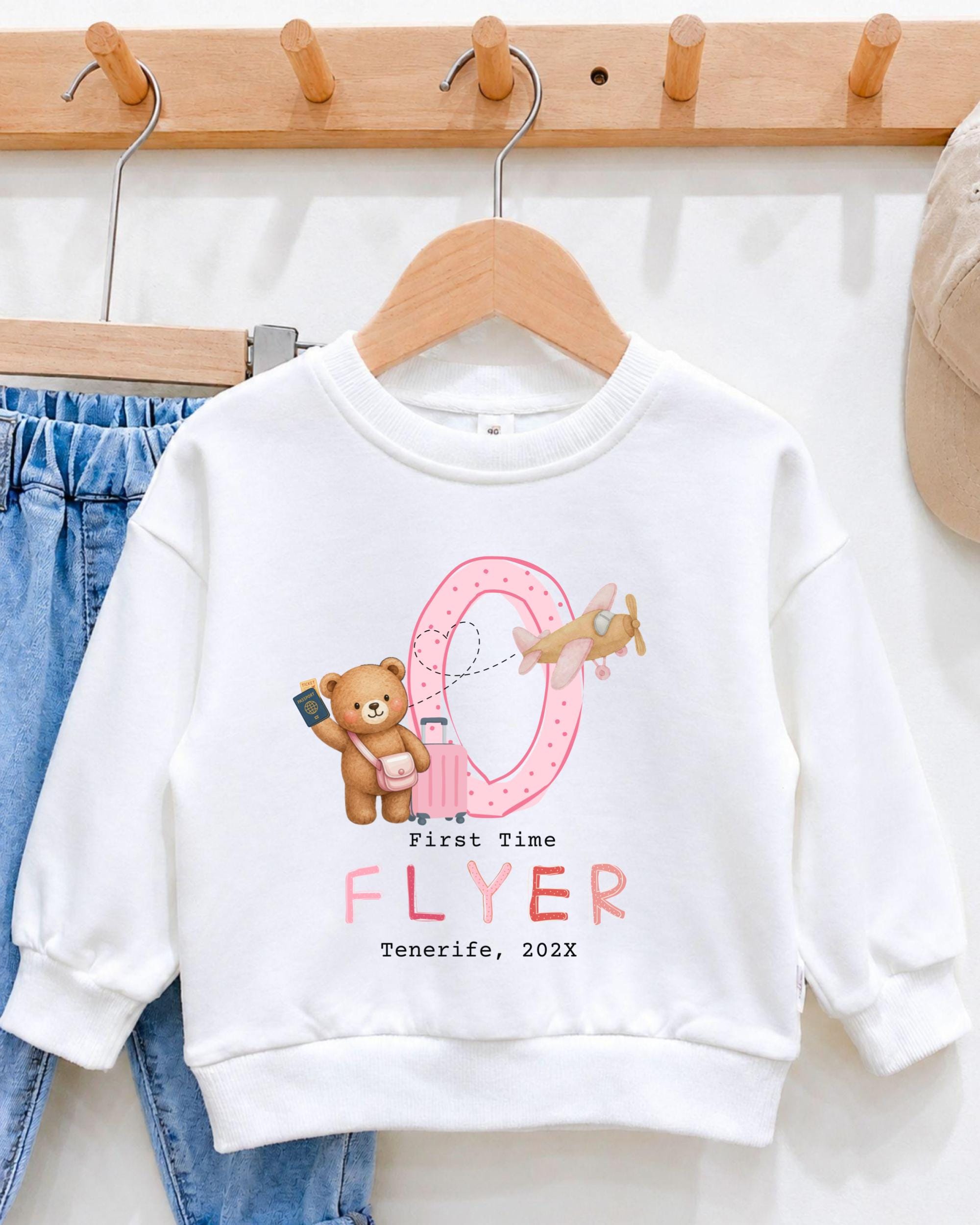 A white sweatshirt with a pink and pink airplane graphic and a teddy bear graphic, along with the text "First Time Flyer" and "Tenerife, 20XX".